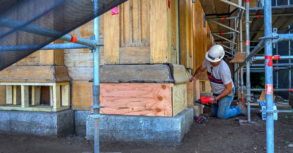 A construction worker repairs wood at the base of Mary Lyon Hall beneath scaffolding during the building’s exterior restoration.