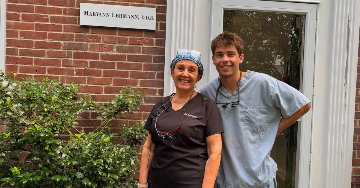 Two smiling individuals stand in front of a brick building, adorned with a sign that reads Maryann Lehmann, DDS. The woman on the right is wearing a surgical cap and a brown shirt. The young man is wearing light blue scrubs.