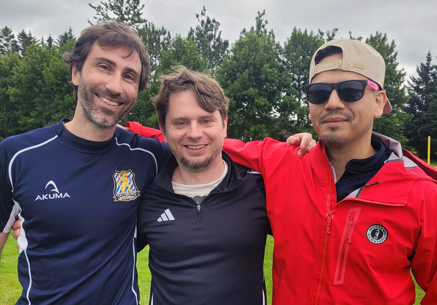 Three men pose together outdoors on a grassy field, smiling at the camera. One wears a navy rugby jersey, another a black Adidas top, and the third a red jacket with sunglasses and a backward cap. Trees and an overcast sky are visible behind them.