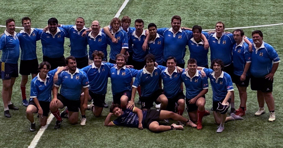A large rugby team in blue jerseys poses for a group photo on an outdoor artificial-turf field. Most players stand in rows while several kneel in front, with one player lying playfully on the ground.
