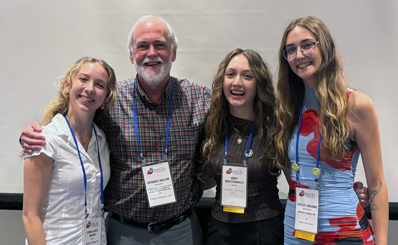 Four individuals smile while standing closely, with their arms behind each others' backs. Three young women and an older man, dressed in business casual attire, each has a nametag lanyard around their neck.