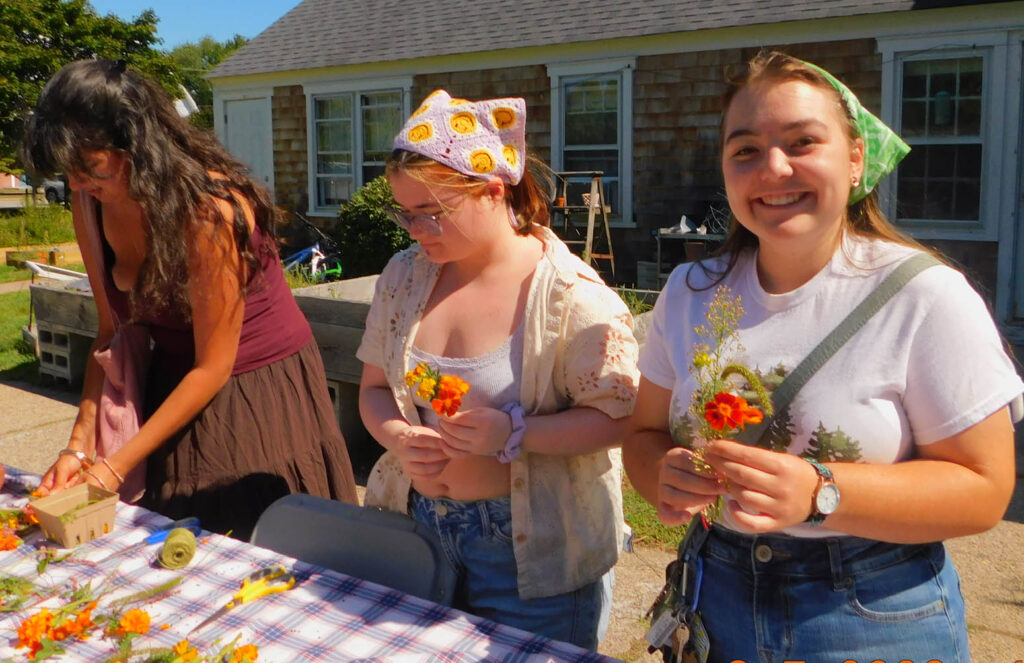 Three young women stand in front of a table with assorted flowers on it. Two of them are holding a small bouquet of flowers in their hands. All are wearing casual outfits while standing outside with a small, single-level house in the background.