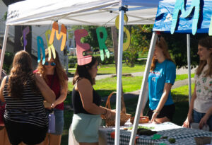 Two young women stand behind a table at an outdoor farmers' market while three female guests talk and explore the items on the table.