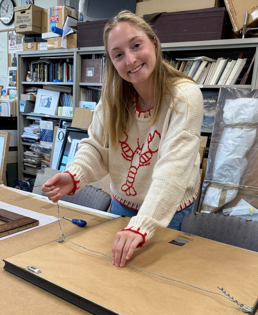 A young smiling woman with long brown hear wearing a beige sweater with a lobster on the front adjust the hanging wire on the back of a large picture frame.