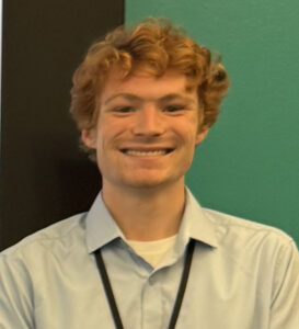 A smiling young man with wavy brown hair wearing a light collared shirt.