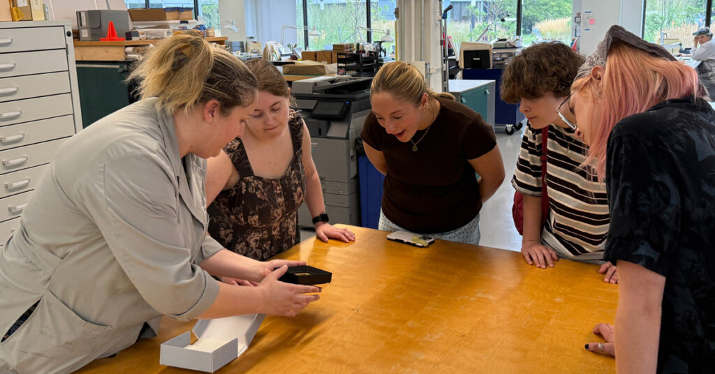 A group of five females gather around a table and look at a small, black rectangular object in an office setting.