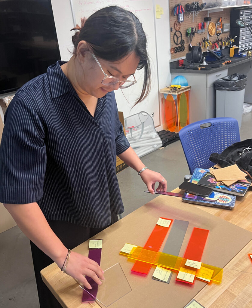 A young woman with dark hear wearing glasses and a dark top concentrates on colorful rectangular swaths while standing at a desk in a classroom.
