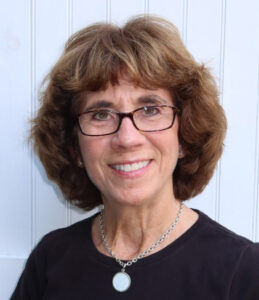 A smiling woman with light brown hair wearing glasses and a black shirt.