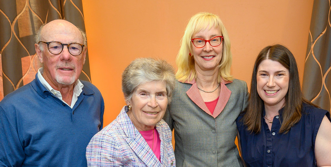 Four individuals, a man and three women, dressed in business attire, smile at an indoor event.
