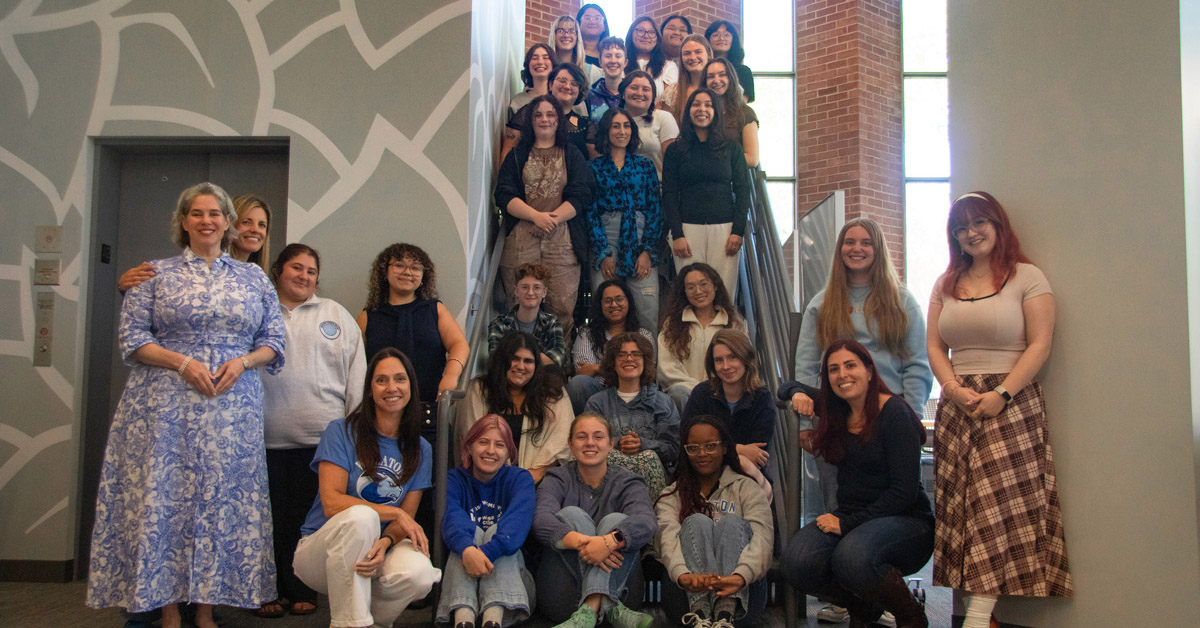 A large group of about 30 women gather on and around an indoor staircase. Most are dressed casually and smiling.