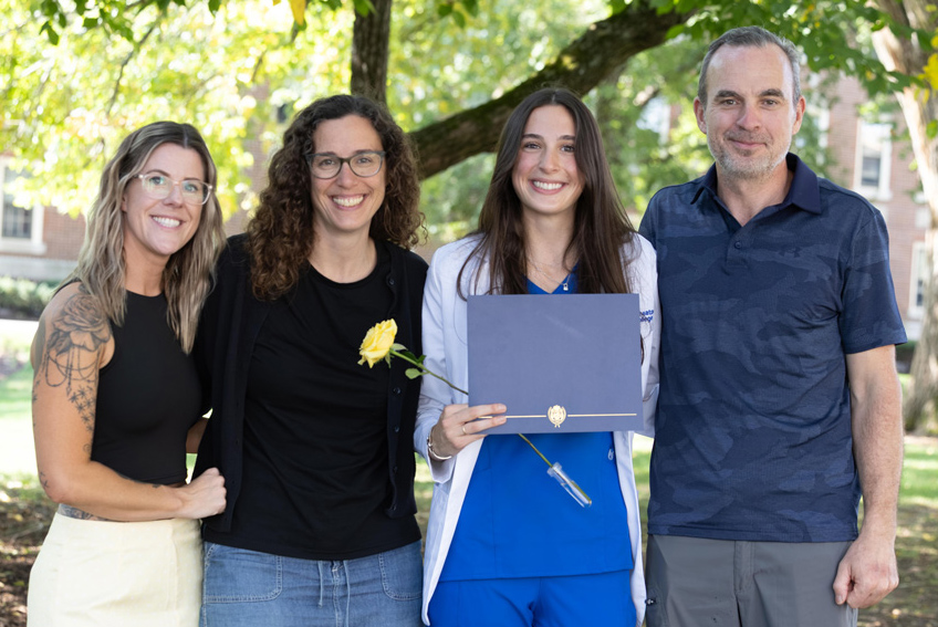 Four smiling individuals stand beside each other. Three older individuals, a man and two women, join a young women with long black hair, wearing a white coat over a blue shirt. She is holding a blueish-gray folder and a yellow rose.