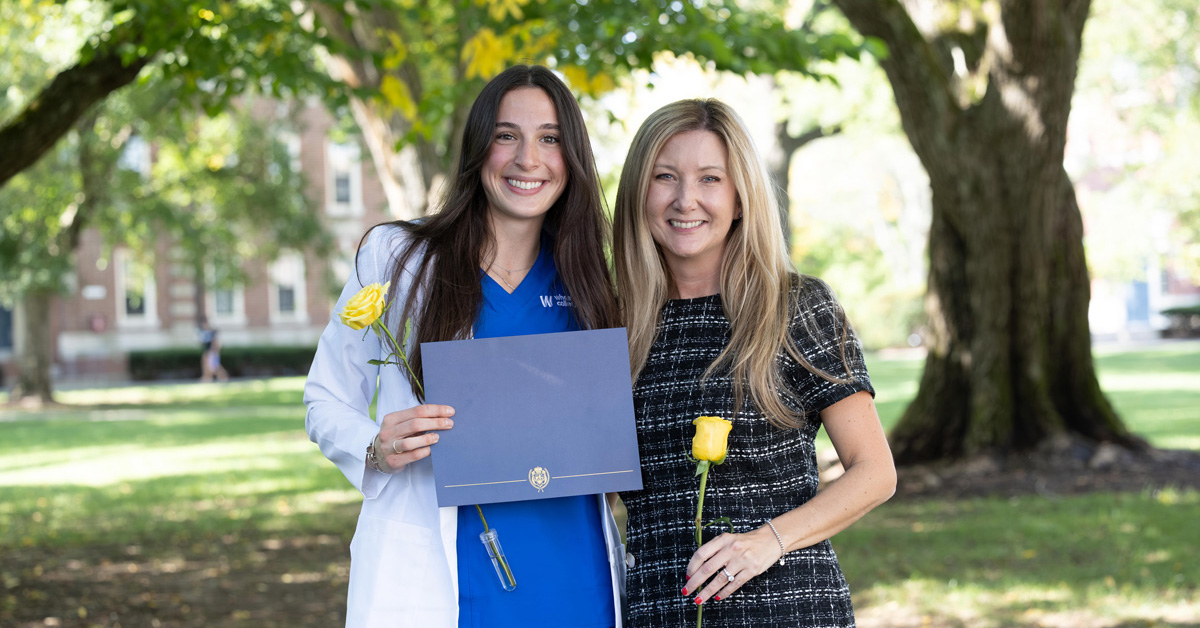 A smiling young woman with long black hair wearing a white coat over a blue shirt holds a greyish-blue folder stands beside a smiling woman with blonde hair wearing a dark dress and holding a yellow rose. Grass, trees with green leaves and brick buildings are in the background.