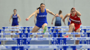 Two young women, one wearing a blue uniform with Wheaton on the shirt and the other wearing a red uniform, leap over hurdles while competing in an indoor race.