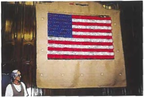 Woven tapestry depiction of an American flag with text incorporated across the stripes, titled "American Families," mounted on a large backing cloth.