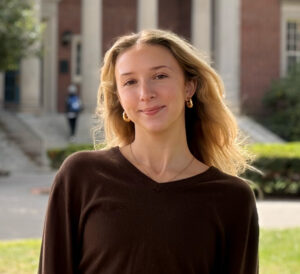 A young woman with long blonde hair wearing a brown shirt stands outside in a grassy area. A brick building with white columns is in the background.