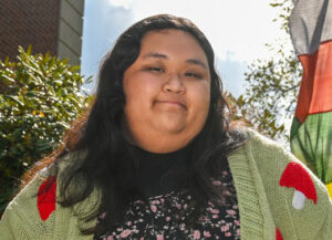 A young woman with long black hair wearing a light green sweater over a dark patterned shirt. Green leaves, blue sky and part of a brick building are in the background.