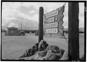 Black-and-white historical photo of the entrance to the Manzanar War Relocation Center, showing a rustic wooden sign mounted on posts with snow-capped mountains in the background.