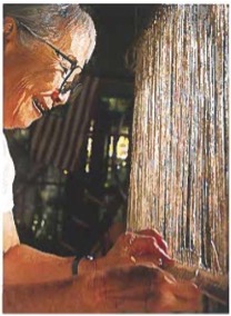 Side profile photograph of artist Momo Nagano smiling while working closely on a weaving loom strung with vertical fibers.