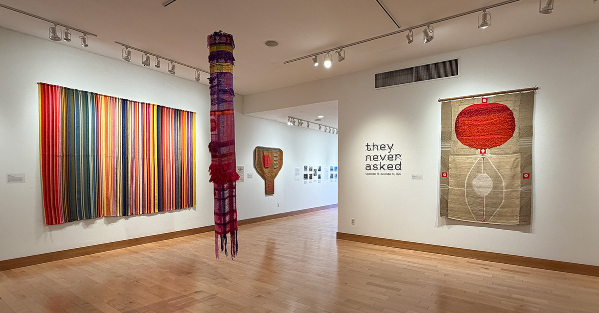 An art gallery exhibition space featuring large colorful woven tapestries, a suspended textile column, and the exhibition title "they never asked" printed on a white wall.